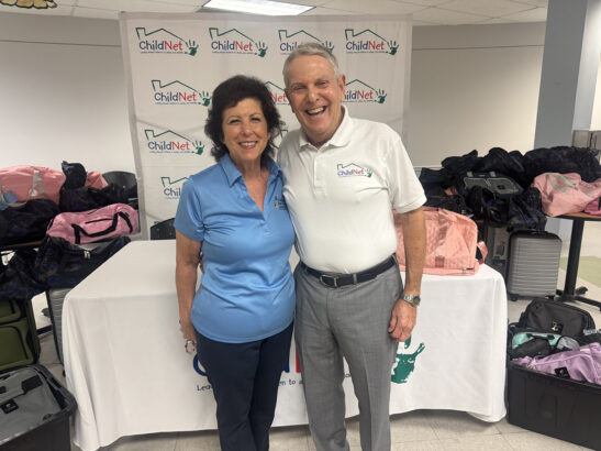 Two smiling adults in ChildNet polo shirts stand by a table with donated duffle bags and a ChildNet banner behind them, appearing to participate in a luggage donation event or gathering. Lifestyle