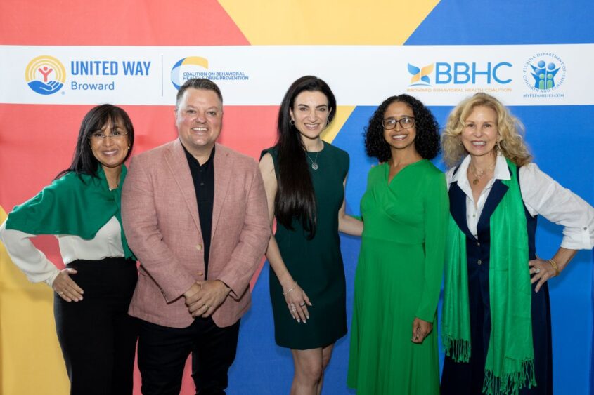 Five people stand smiling in front of a colorful backdrop featuring logos for United Way Broward, BBHC, and behavioral health organizations. Four women and one man are dressed in business casual attire. Lifestyle