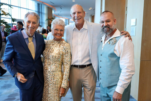 Four smiling adults, dressed formally, pose together at the Broward Partnership’s annual event. The group, attending the Salute to Leadership, stands before bright windows with other guests mingling in the background. Lifestyle
