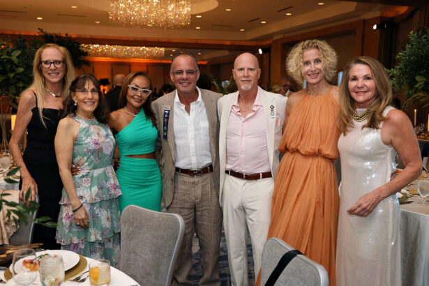 Seven adults dressed in formal attire stand together, smiling for a group photo at the Annual Broward Partnership Salute to Leadership event, with chandeliers, greenery, and set dining tables creating an elegant backdrop. Lifestyle