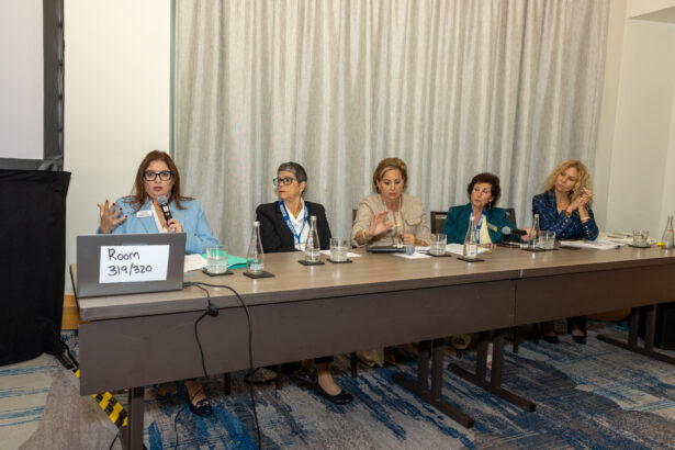 Five women sit at a conference table during a panel discussion. One woman speaks into a microphone, while the others listen. A laptop with a sign reading "Room 319/320" is on the table. The background features curtains. Lifestyle