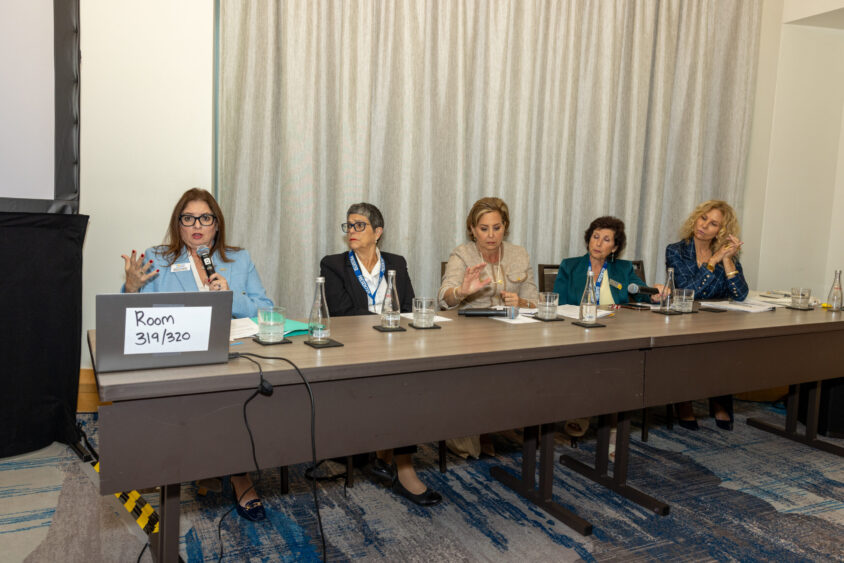 P3. Monica Cepero, Silvia Quintana, Jennifer O'Flannery, Cindy Arenberg Seltzer, and Kathleen Cannon Five women sit at a conference table during a panel discussion. One woman speaks into a microphone, while the others listen. A laptop with a sign reading "Room 319/320" is on the table. The background features curtains. Lifestyle