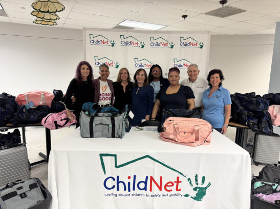 A group of seven adults stands behind a table covered with duffel bags for a ChildNet duffle bag donation. More bags and suitcases are in the background, and everyone appears smiling in the well-lit indoor setting. Lifestyle