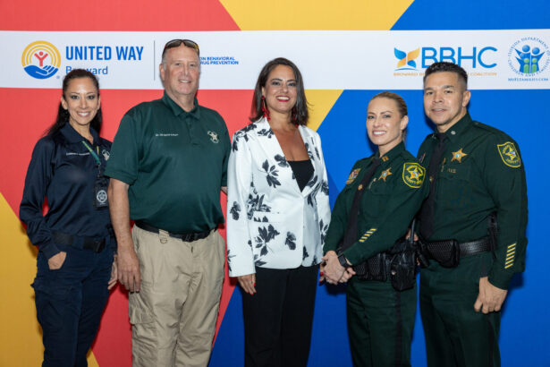 Five people stand smiling in front of a backdrop with United Way and other logos. Two women and three men, some in law enforcement uniforms, pose for a group photo at an event. Lifestyle