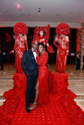 A couple dressed formally poses on a path of red roses at the Mayors’ Gala, with three performers in elaborate rose costumes standing behind them in a glamorous indoor setting supporting United Way Broward housing initiatives. Lifestyle