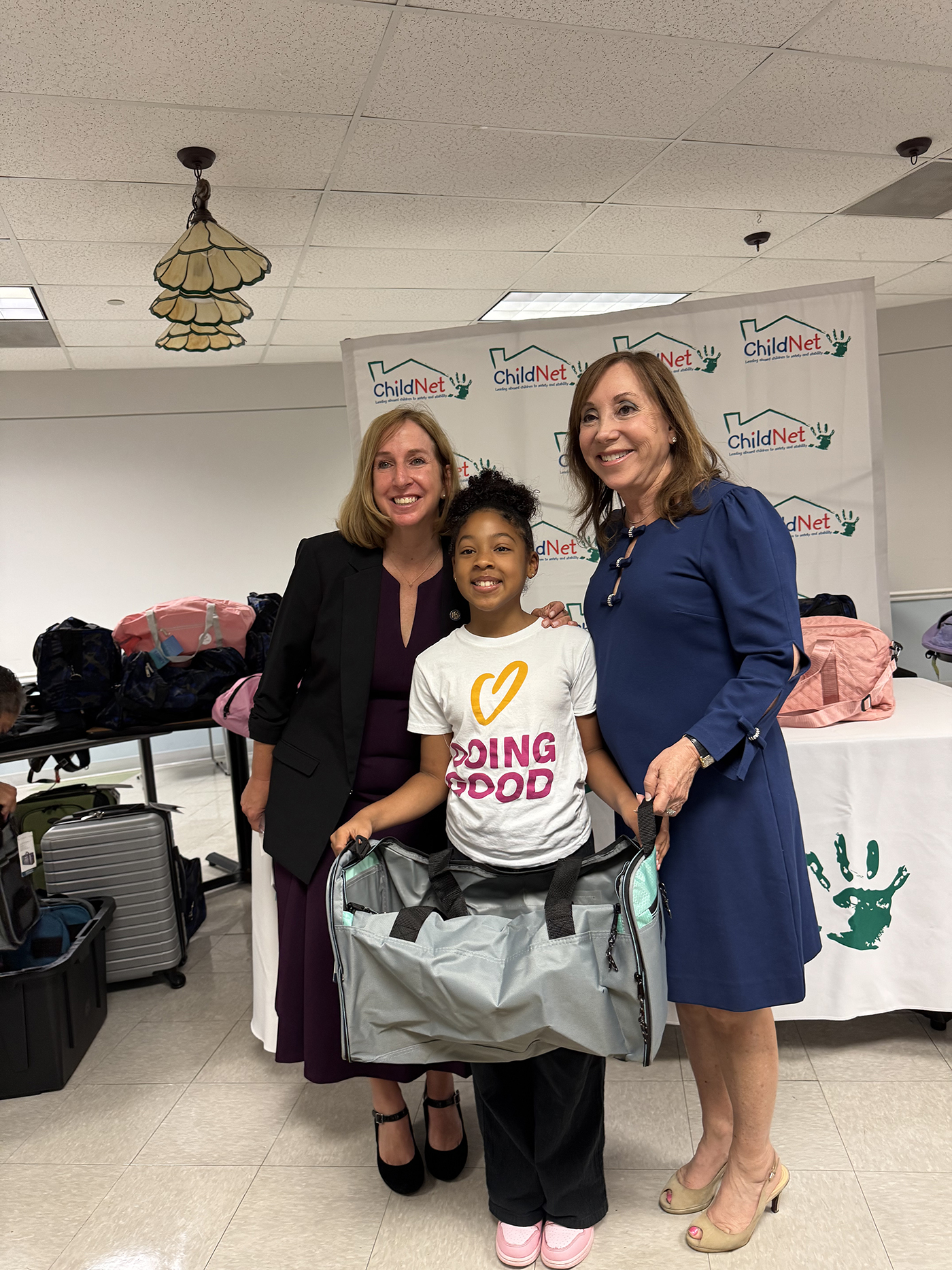 Three women smile together as the young girl in the center holds an open duffle bag, wearing a "Doing Good" T-shirt. With ChildNet banners and luggage donation items behind them, they show their support for children in need. Lifestyle