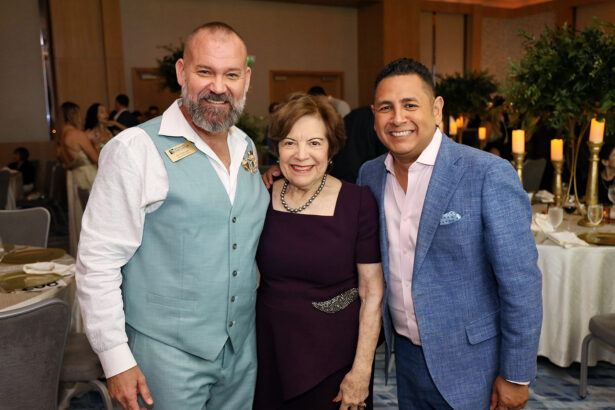 Three people dressed in formal attire smile and pose together at the Broward Partnership's Annual event, Salute to Leadership, with decorated tables and greenery in the background. Lifestyle