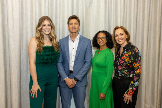 Four people stand side by side in front of a light-colored curtain, dressed in semi-formal attire: two women in green outfits, a man in a blue suit, and a woman in a floral blouse and black pants. All are smiling at the camera. Lifestyle