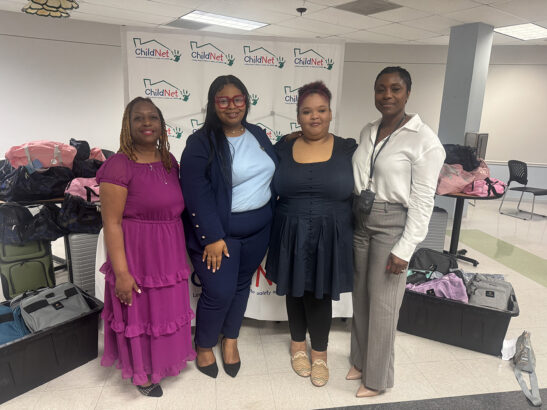 Four women stand side by side, smiling in front of a ChildNet banner. Tables behind them display bags and supplies, highlighting a Luggage Donation drive at this indoor event or gathering. Lifestyle