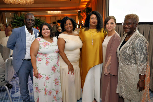 Six people dressed in formal attire stand together and smile for a group photo at the Broward Partnership's Salute to Leadership annual event, with warm lighting and decorated tables in the background. Lifestyle