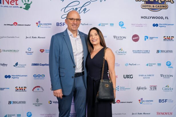 A man in a blue suit and a woman in a black dress pose together, smiling, in front of a step-and-repeat banner with sponsor logos at the ChildNet Hosts Inaugural Ripple Effect Gala formal event. Lifestyle
