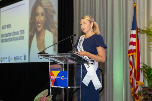 A woman in a navy dress and sash reading "Miss Southern Shores" speaks at a podium. Behind her is a large projected photo of her and a U.S. flag. She appears to be at a formal event or conference. Lifestyle