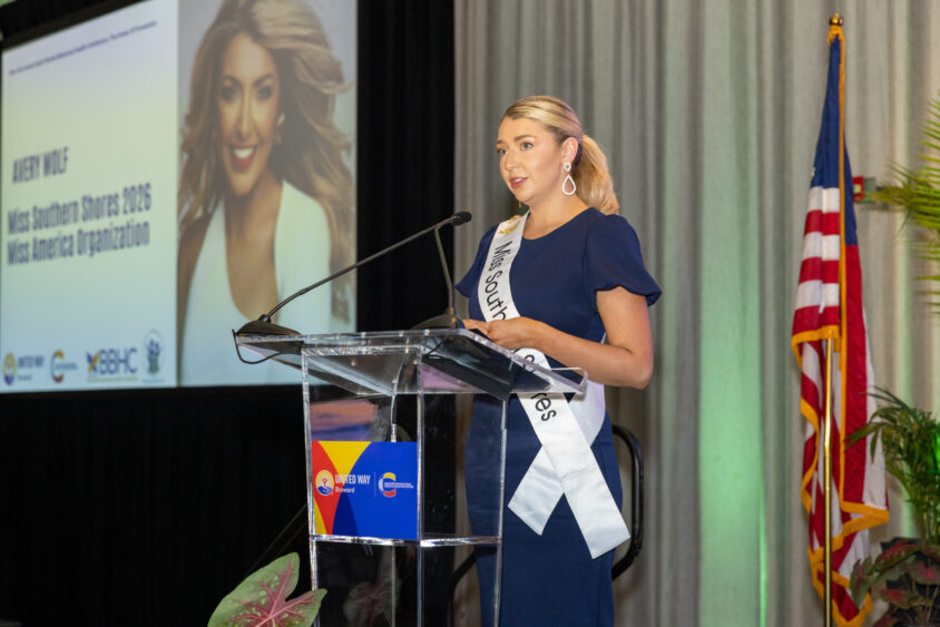P6. Avery Wolf A woman in a navy dress and sash reading "Miss Southern Shores" speaks at a podium. Behind her is a large projected photo of her and a U.S. flag. She appears to be at a formal event or conference. Lifestyle