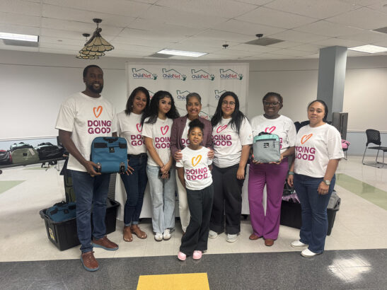 A diverse group of eight people, including a child, stand indoors wearing “Doing Good” shirts. They smile at the camera, holding duffle bags and supplies, with bins and a “ChildNet” banner in the background, supporting luggage donation. Lifestyle