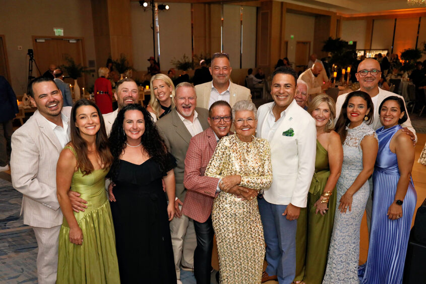 A group of people dressed in formal and semi-formal attire stand close together, smiling for a group photo at Broward Partnership’s annual Salute to Leadership event, with decorated tables visible in the background. Lifestyle