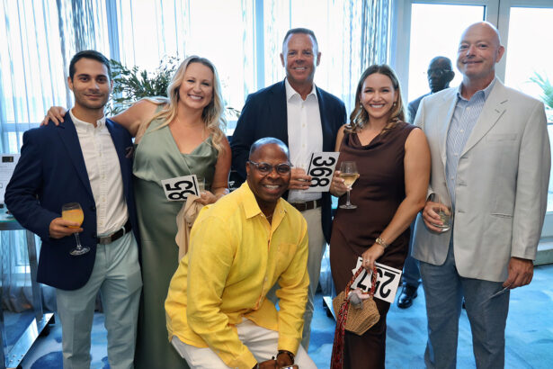 A group of six adults, dressed in formal attire, smile and pose together at an indoor Broward Partnership event. Some hold drinks and numbered cards, suggesting the Annual Salute to Leadership fundraiser. Light filters through large windows behind them. Lifestyle