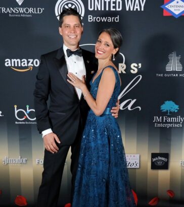 A smiling couple poses on the red carpet at the Mayors’ Gala, a formal event supporting United Way Broward housing initiatives. The man wears a black tuxedo while the woman stuns in a sleeveless blue gown, with sponsor logos displayed behind them. Lifestyle
