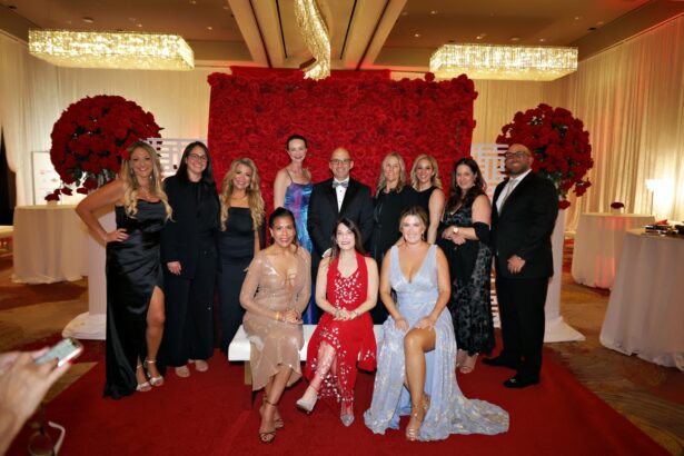 A group of eleven people dressed in formal evening attire pose together in front of a wall of red roses at the glamorous Mayors’ Gala, an indoor event supporting United Way Broward and local housing initiatives, with elegant lighting and decor. Lifestyle