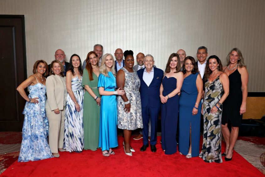 A group of people in formal attire stand together on a red carpet in front of a beige wall, smiling for a group photo at the Ripple Effect Gala hosted by ChildNet. Lifestyle