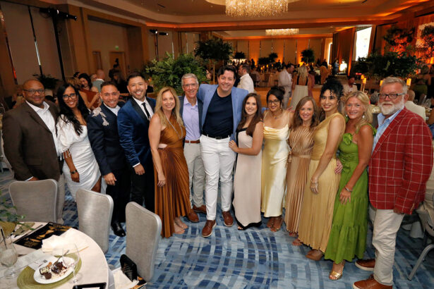 A group of people dressed in formal attire stand together, smiling for a photo at Broward Partnership’s Annual Salute to Leadership banquet in a large, elegantly decorated room with round tables and a chandelier overhead. Lifestyle