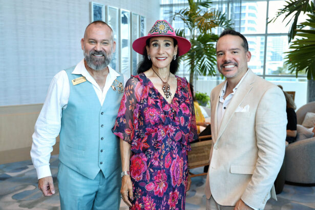 Three people smile at the camera during the Broward Partnership Annual Leadership Event. The man on the left wears a light blue vest, the woman in the center dons a floral dress and hat, and the man on the right is in a beige suit. Plants fill the background. Lifestyle