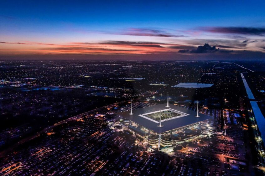 Aerial view of a brightly lit World Cup stadium surrounded by packed parking lots at dusk, with city lights spreading under a colorful sunset sky and water visible in the distance. Lifestyle