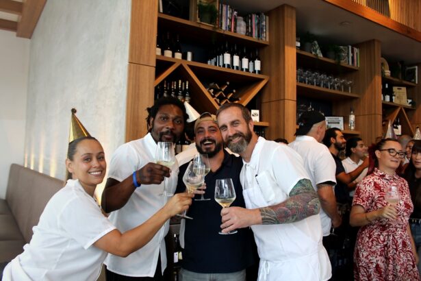 Four people, three in white outfits, smile and raise wine glasses in a lively bar or restaurant as Ukiah celebrates its first anniversary. Shelves of wine bottles and glasses line the background while other guests mingle behind them. Lifestyle