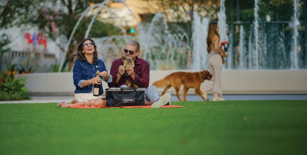 A smiling couple sits on a red blanket in an inclusive park near a fountain, opening a bottle and holding a small dog. In the background, a woman walks a large brown dog amid trees and water spray. Lifestyle