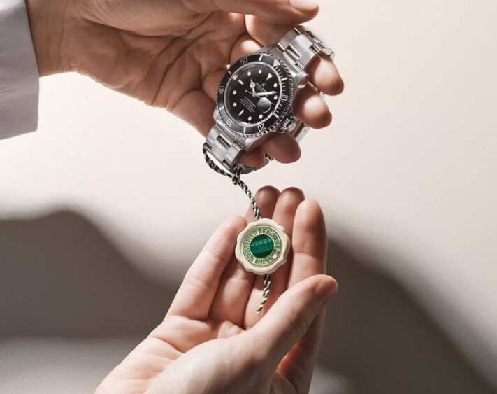 A person holds a silver Rolex watch in one hand and a green Rolex certification tag in the other, against a neutral background—an elegant display of Weston Jewelers' Rolex Certified Pre-Owned collection. Lifestyle