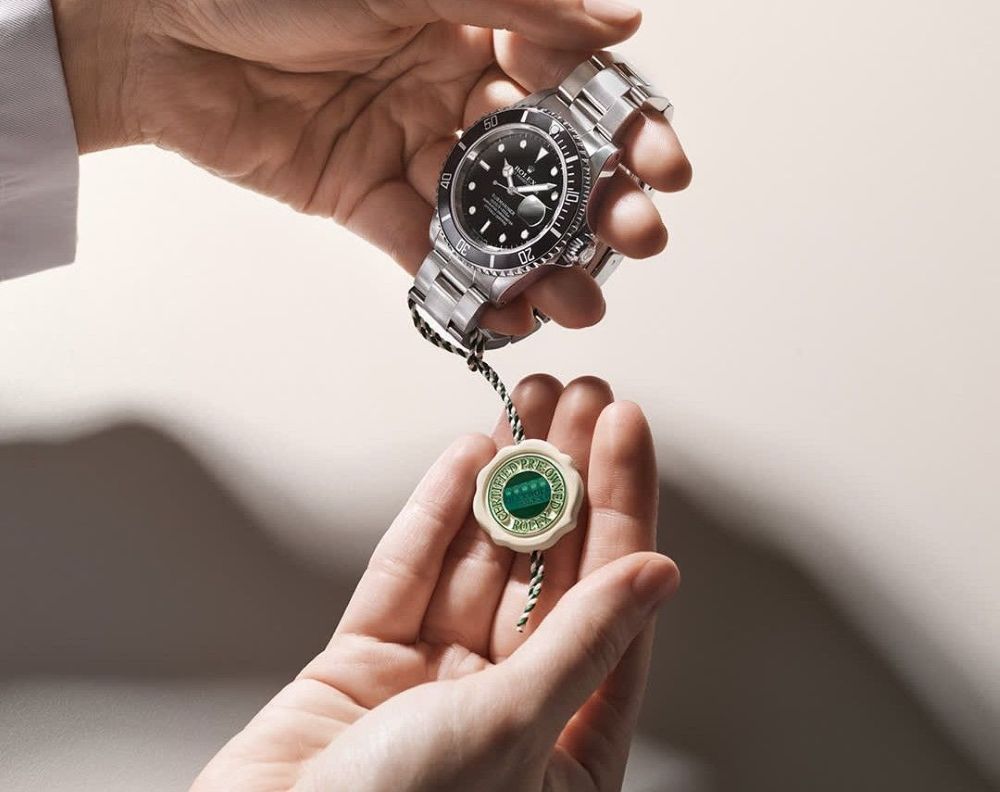 A person holds a silver Rolex watch in one hand and a green Rolex certification tag in the other, against a neutral background—an elegant display of Weston Jewelers' Rolex Certified Pre-Owned collection. Lifestyle