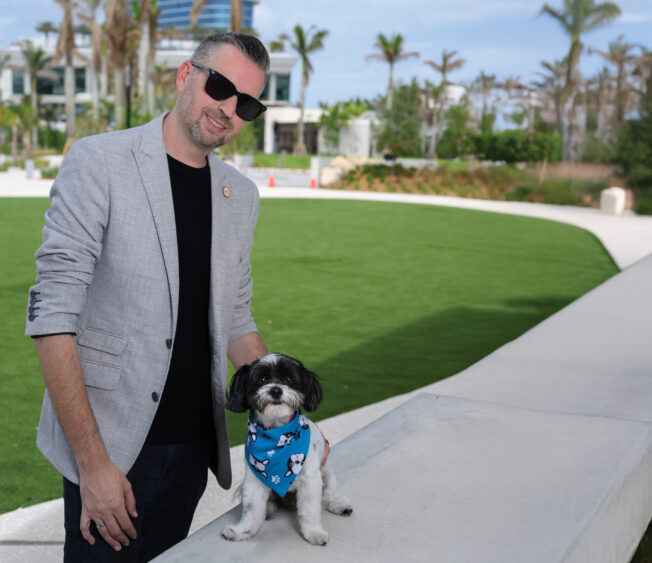 A man in a gray blazer and sunglasses stands outside by a green lawn, smiling and petting a small black and white dog wearing a blue bandana, enjoying the Dog Days of Summer. Lifestyle