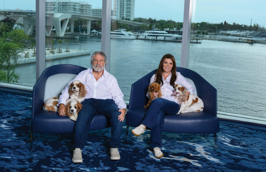 Susan Renneisen and her companion sit on blue chairs by large windows overlooking the water, each holding two small dogs in their laps. Dressed alike in white tops and jeans, they enjoy these Dog Days of Summer with their furry friends, while boats drift past outside. Lifestyle