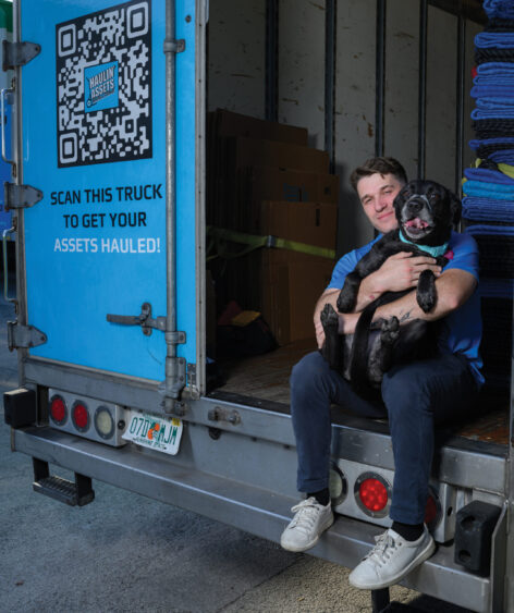 A man sits on the back of a moving truck, smiling and holding a happy black dog—Gizmo—enjoying the Dog Days of Summer. The truck door displays a QR code, the company logo, and the text "Scan this truck to get your assets hauled!. Lifestyle