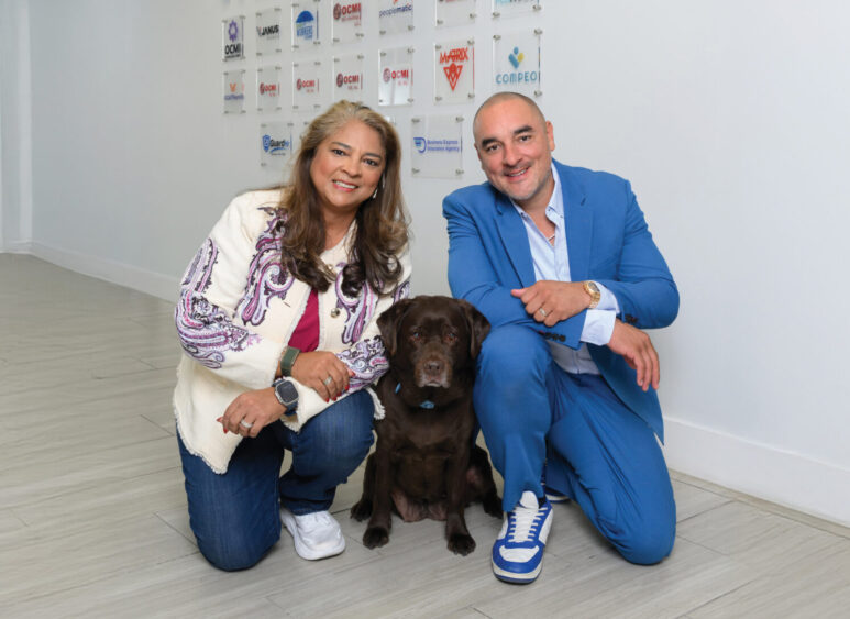 Oscar Montenegro and Melanie Montenegro kneel beside a brown dog in a hallway with light wood flooring, smiling at the camera during the Dog Days of Summer. Behind them, a wall displays the words "WE ARE ONE" and several business logos. Lifestyle