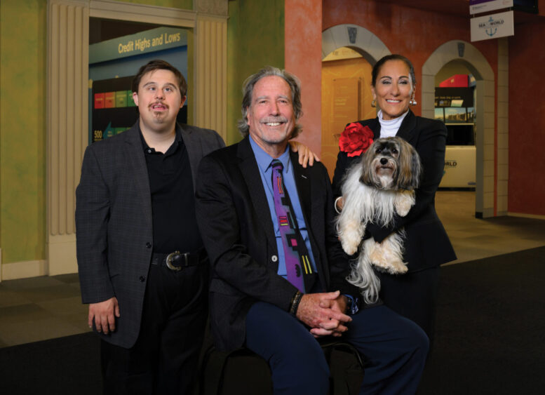 Three people, one seated and two standing, pose indoors with a small dog at what appears to be a Dog Days of Summer event. The setting resembles a lobby or public space, with columns and signs in the background. All are smiling and well-dressed—perhaps gathering to meet Jackie Ducci or celebrate the occasion together. Lifestyle