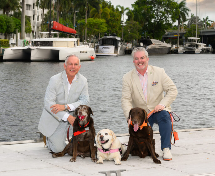 Two men in light suits, Bob Swindell and John Cotter, kneel on a dock by the water with three dogs—two brown and one white—enjoying the Dog Days of Summer. Boats and palm trees are visible in the background on a cloudy day. Lifestyle