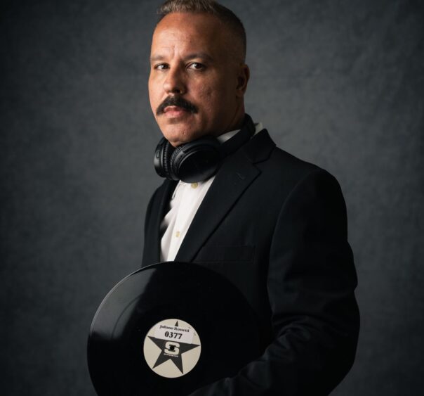 A man in a black suit with headphones around his neck holds a black vinyl record, ready to match the perfect music to the moment. He has short hair, a mustache, and stands against a dark textured background. Lifestyle