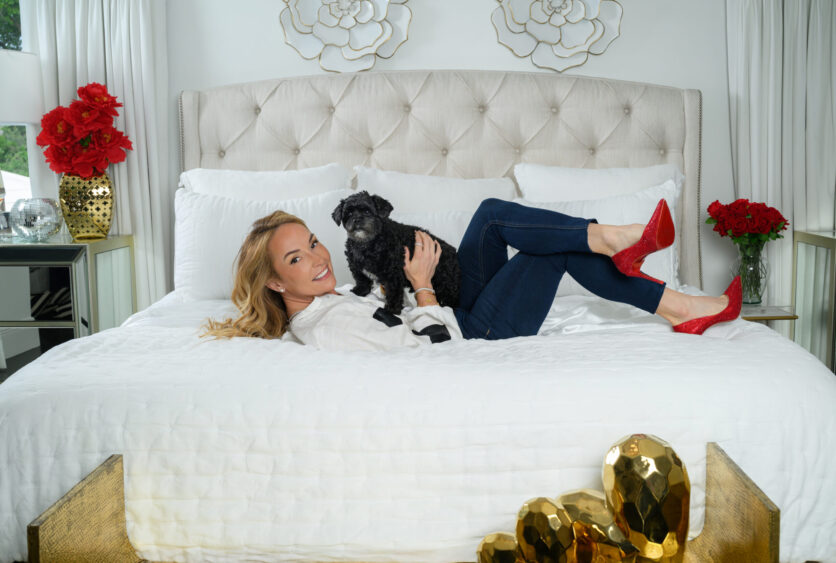 A woman in red heels lies on a white bed, smiling and holding a small black dog during the Dog Days of Summer. Gold decorative objects are at the foot of the bed, with red flowers on bedside tables—a scene inspired by Daren Koenig Cronin. Lifestyle