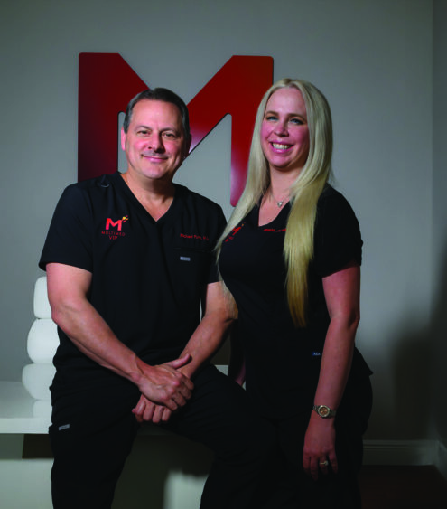 Two people in black outfits stand side by side, smiling, in a modern room with a wooden floor—a space that radiates both fitness and beauty. A large red "M" logo is displayed on the wall behind them. Lifestyle