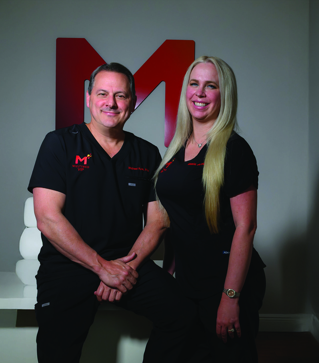 Two people in black outfits stand side by side, smiling, in a modern room with a wooden floor—a space that radiates both fitness and beauty. A large red "M" logo is displayed on the wall behind them. Lifestyle