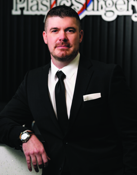 A man in a black suit and tie stands confidently indoors, resting his hand on a counter. He has short hair and a trimmed beard. A sign for a plastic surgery office—emphasizing beauty and health care—is visible in the background. Lifestyle