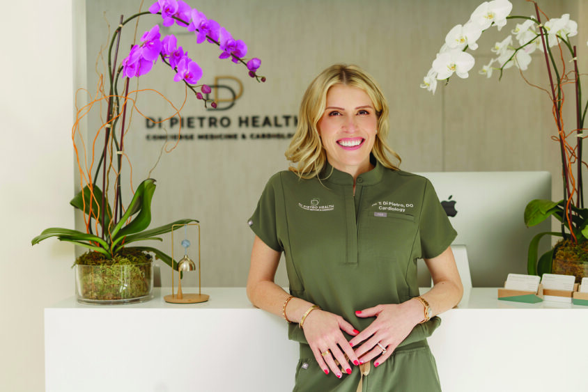 A smiling woman in olive scrubs stands at a reception desk with orchids, a computer, and business cards. The sign behind her reads “DIPietro Health.” The setting appears to be a modern medical office dedicated to fitness and health care. Lifestyle