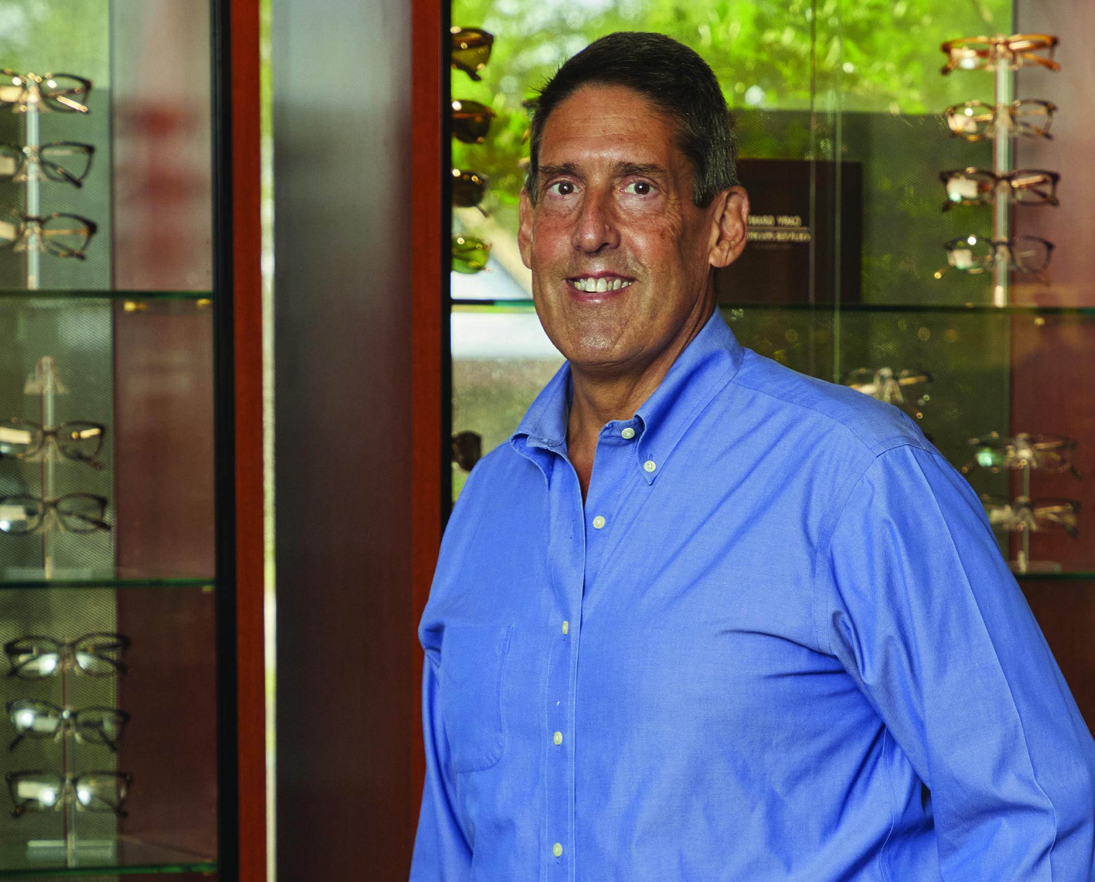 A man in a blue shirt smiles while standing in front of glass display cases filled with eyeglasses, radiating confidence and well-being—a perfect blend of health care and beauty—with a green, leafy background outside the window. Lifestyle