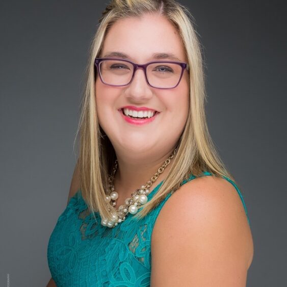 Smiling woman with blonde hair, wearing glasses, a teal lace top, and a chunky pearl necklace from Daoud’s Fine Jewelry, posed against a plain gray background. Lifestyle