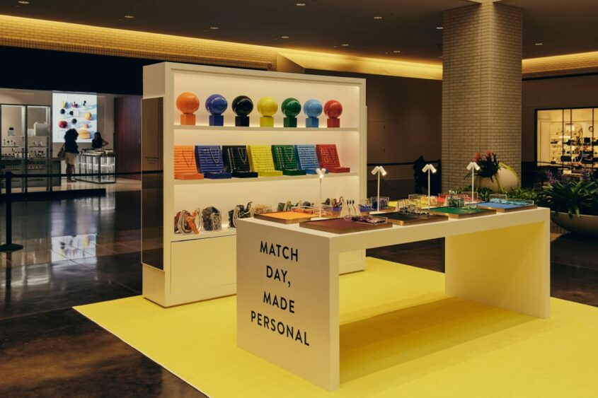 A modern display in Aventura Mall features colorful books, round objects, gift items from the Kendra Scott Color Bar, and a white table with “MATCH DAY, MADE PERSONAL” printed on it, all set on a bright yellow rug. Lifestyle