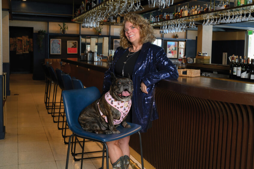 A woman in a blue sequin blazer stands by a bar, smiling, with a brindle French bulldog in a pink harness sitting on a barstool next to her. Bottles and glasses line the bar in the background, capturing the spirit of Dog Days of Summer as celebrated by Laine Doss. Lifestyle