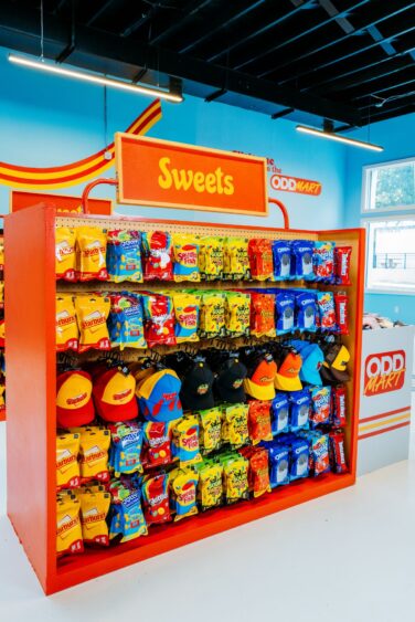 Bright orange display shelf labeled "Sweets" holds various colorful candy bags and hats in a Po-Up Run ODDMART store with blue walls and large windows. The display is neatly organized, eye-catching, and perfect for spotting limited-time closes out treats. Lifestyle
