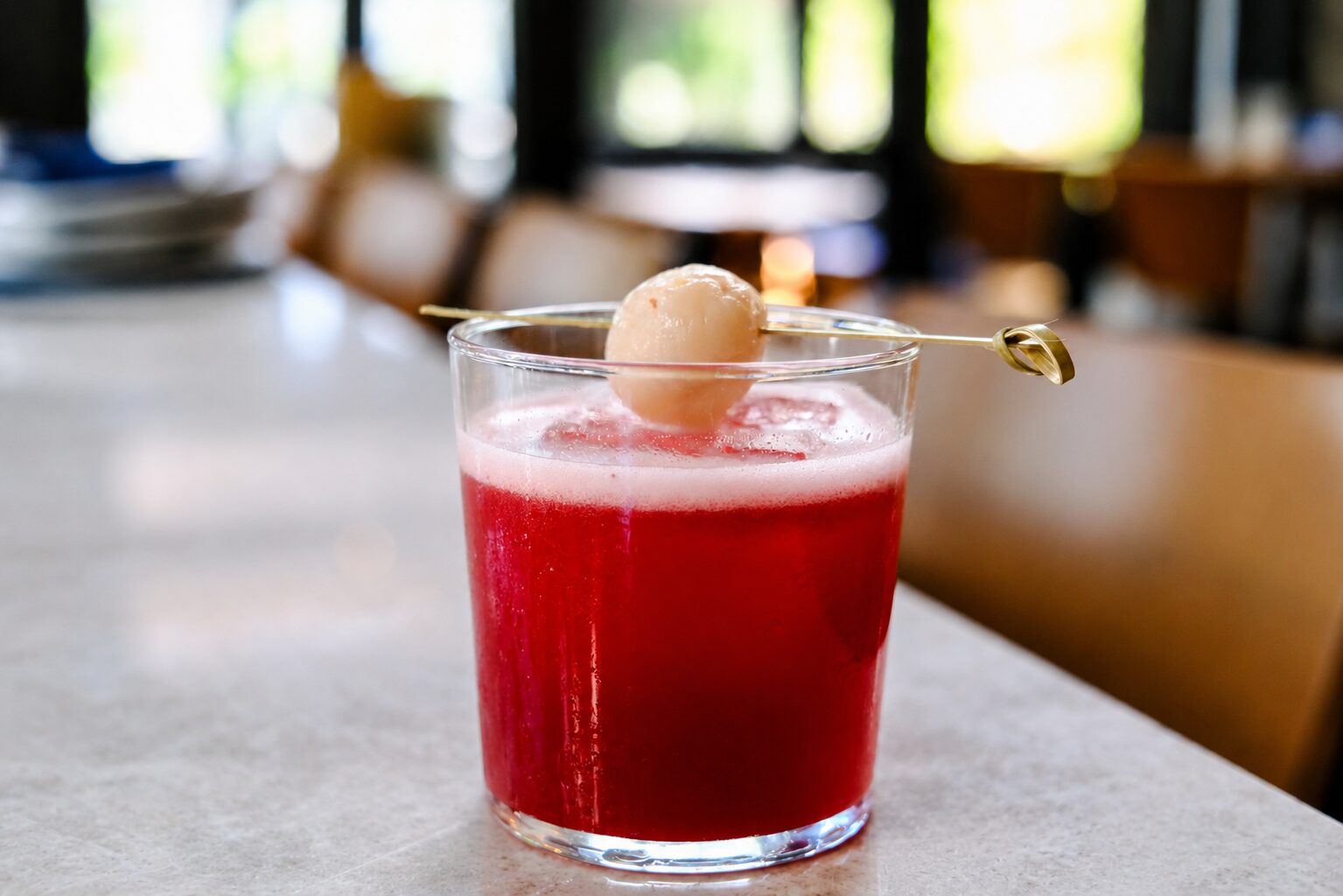 A red Pear Necessities cocktail in a glass, topped with foam and garnished with a lychee on a cocktail pick, sits on a light-colored surface with blurred chairs and windows in the background—a drink crafted for a good cause. Lifestyle