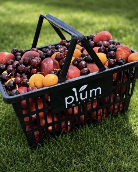 A black basket filled with cherries, apricots, peaches, and nectarines sits on green grass. The basket has a "plum market" logo on the side. Lifestyle