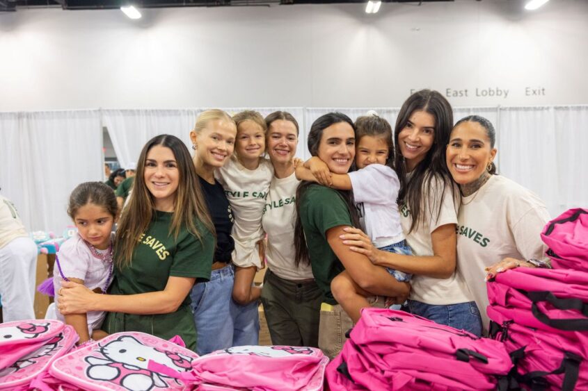 Eight smiling women and girls stand behind a table with pink Hello Kitty backpacks, posing for a group photo at an indoor Back-to-School event in Miami Beach. Most wear green or white shirts. White curtains and signs are visible in the background. Lifestyle
